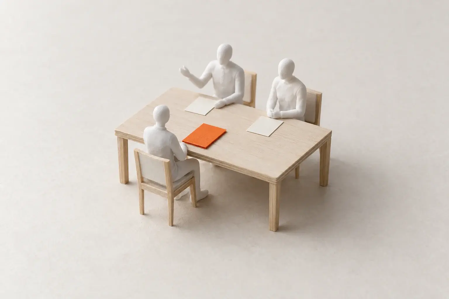 A paper model of two figures across a table from a candidate, with an orange folder in front of the candidate.