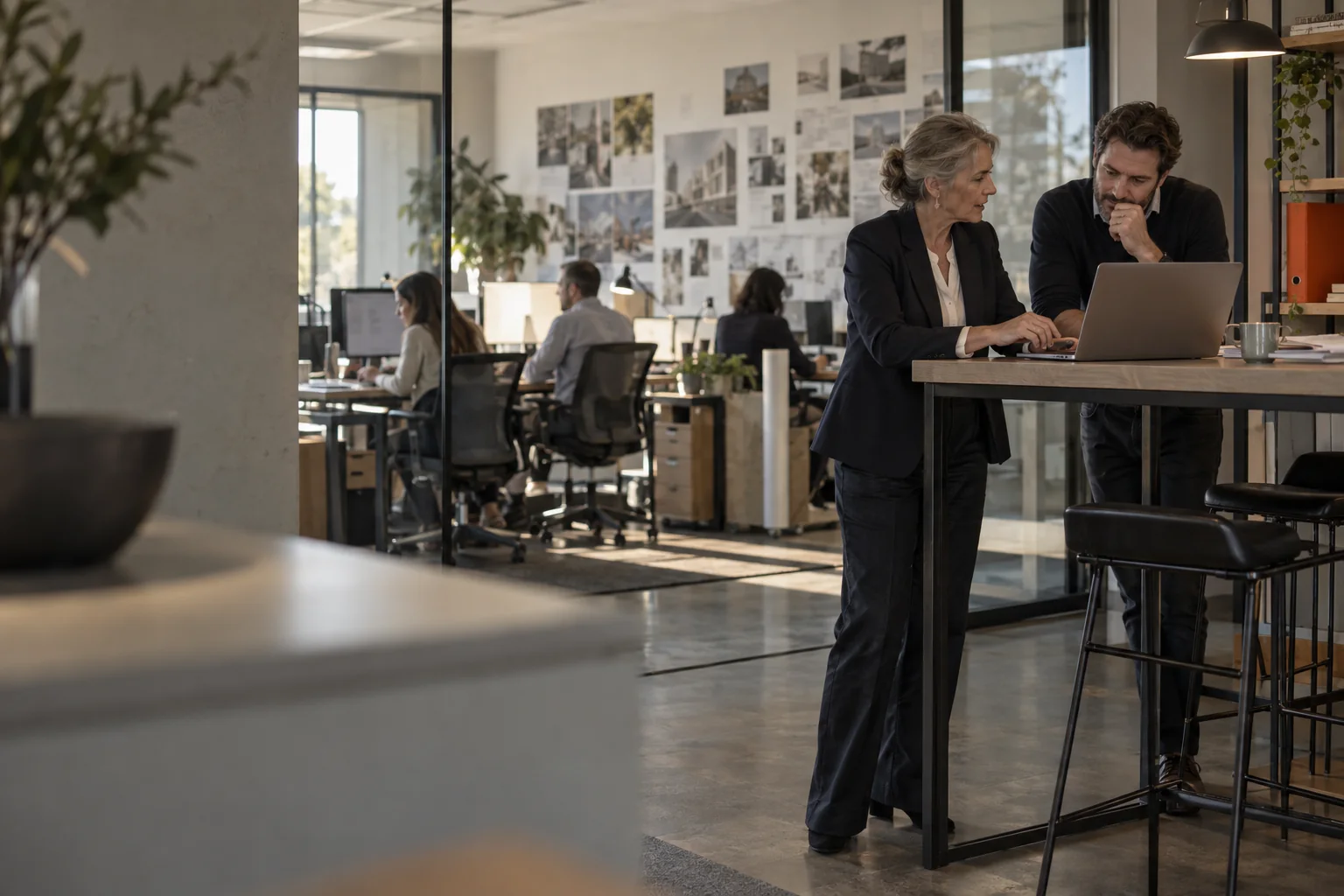 An agency's managing partner and operations director talk over a laptop at a high counter, with the studio behind a glass wall.